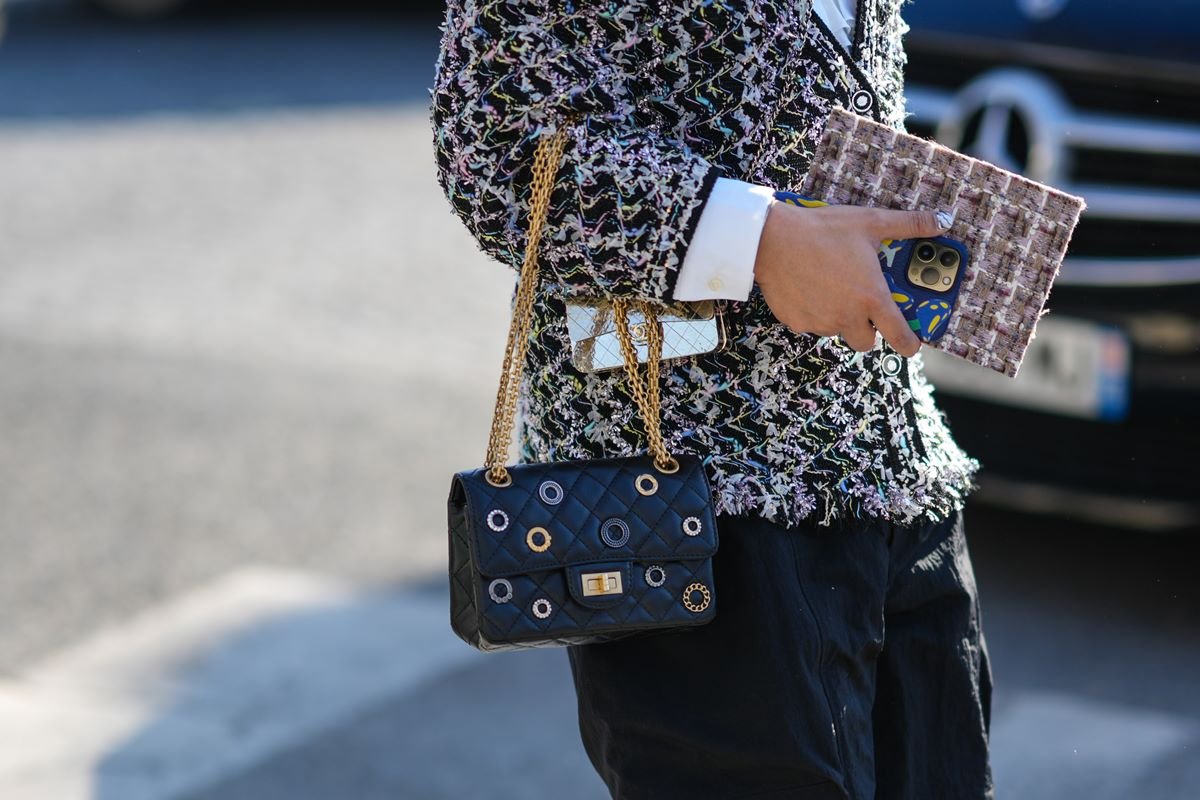 Uma convidada usa uma jaqueta de tweed com estampa xadrez preto e branco, uma micro bolsa dourada brilhante metálica da Chanel, uma bolsa preta brilhante de couro com tachas prata e ouro 2.55 da Chanel, shorts pretos, fora da Chanel, durante a Semana de Moda de Paris - Moda Feminina F/ W 2022-2023, em 08 de março de 2022 em Paris, França. - Metrópoles