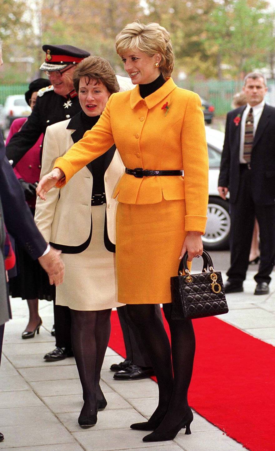Princesa Diana, uma mulher jovem, branca e com cabelo loiro curto, comprimentando pessoas. Ela usa um conjunto laranja de blazer e saia, meia calça preta e uma bolsa preta da Dior. - Metrópoles