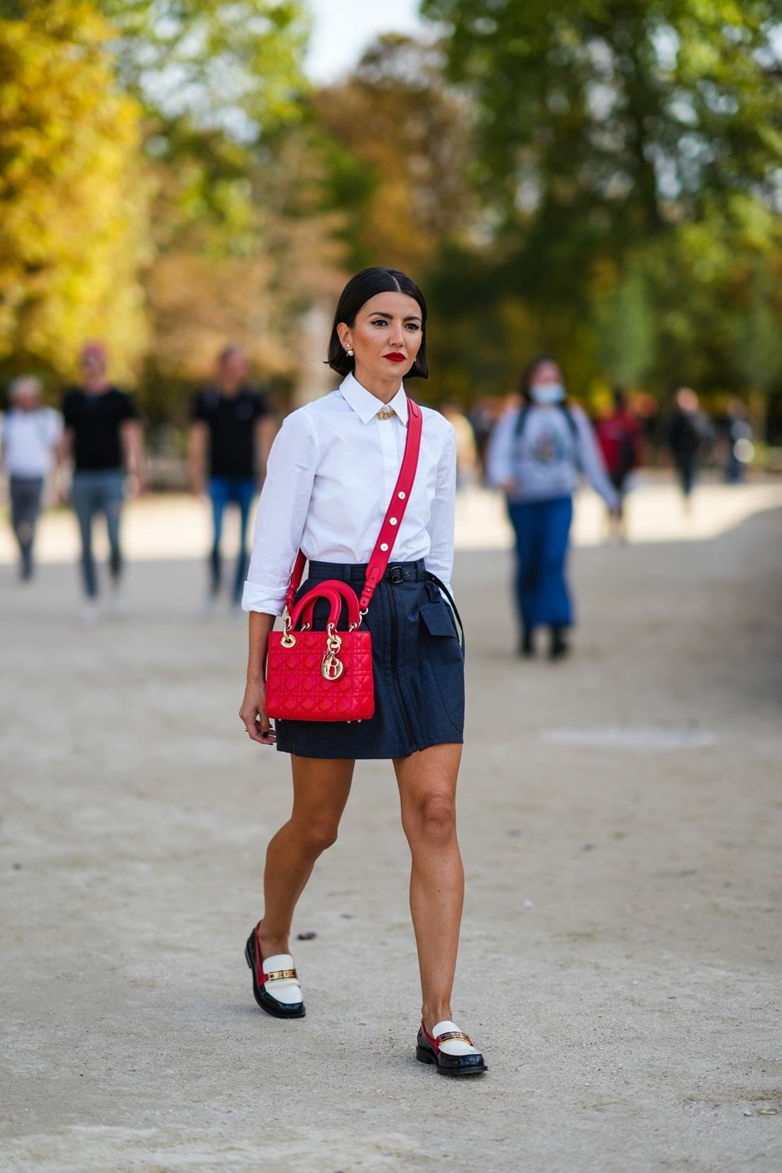 Uma mulher branca e jovem, de cabelo preto curto e liso, anda pelas ruas de Paris durante a Semana de Moda em 2021. Ela usa uma camisa branca de botão, uma saia preta curta, uma bolsa vermelha modelo Lady Dior, da marca Dior, e um sapato mocassim de couro preto e branco. - Metrópoles