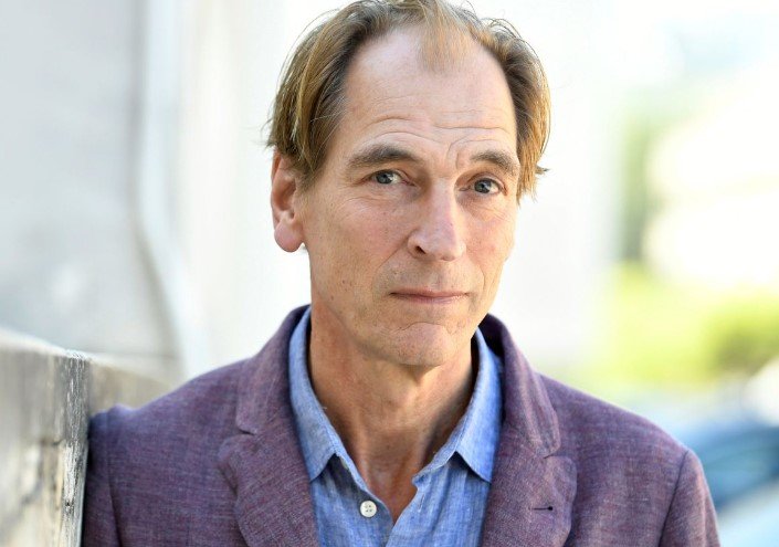 Foto colorida do ator Julian Sands. Ele está de terno roxo e camisa azul - Metrópoles