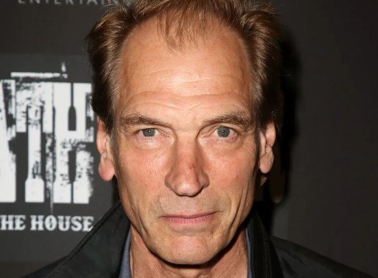 Foto colorida do ator Julian Sands. Ele está de roupa escura - Metrópoles