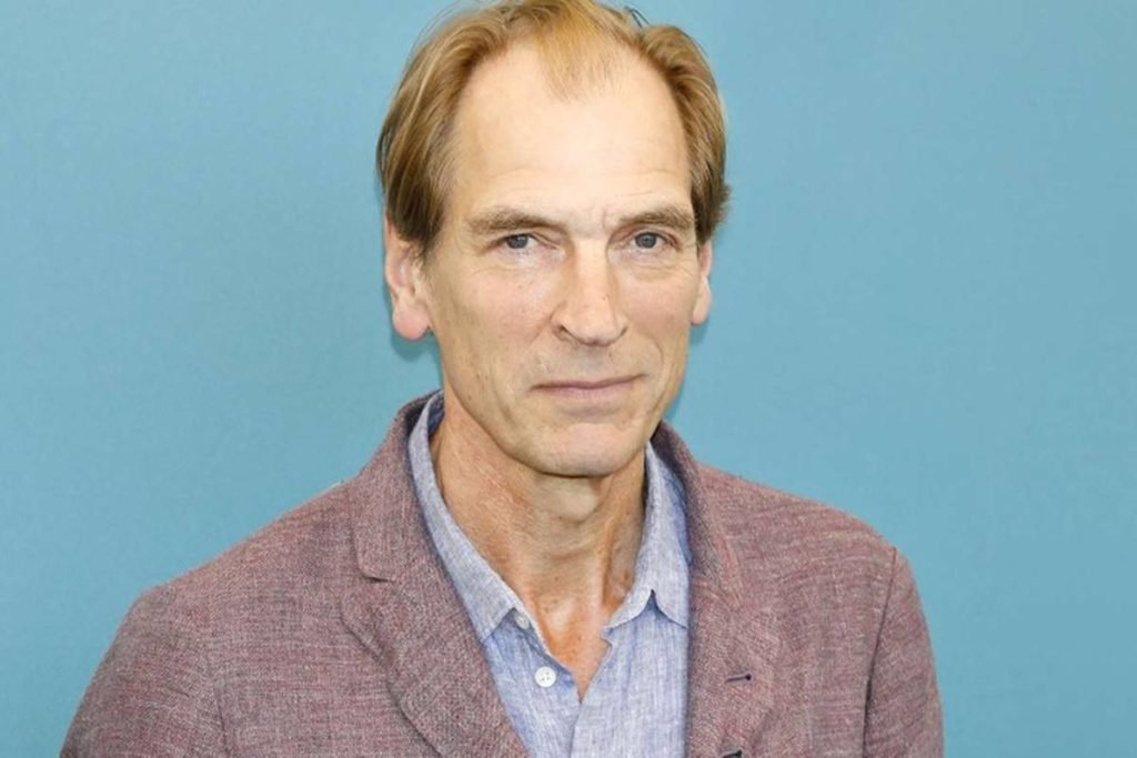Foto colorida do ator Julian Sands. Ele está de terno roxo e camisa azul - Metrópoles