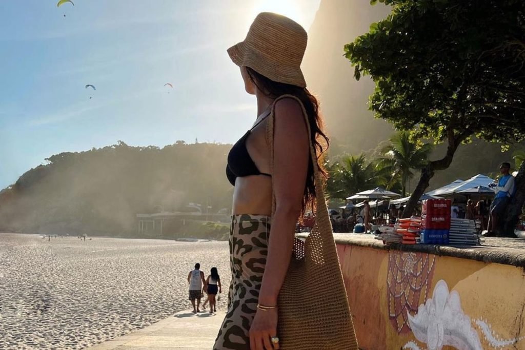 Mulher na praia, de lado. Ela usa chapéu e bolsa de palha, além de bíquini preto e calça de animal print - Metrópoles