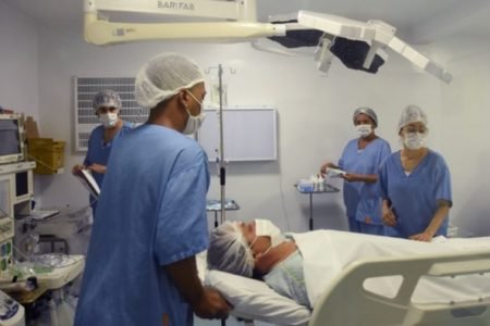 Imagem colorida de uma sala de cirurgia onde há profissionais da saúde vestindo roupas hospitalares na cor azul