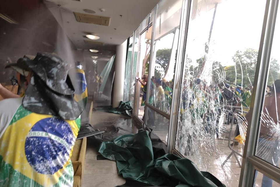 foto colorida do prédio do STF com vidros quebrados. Homem com camisa do Brasil e manifestantes