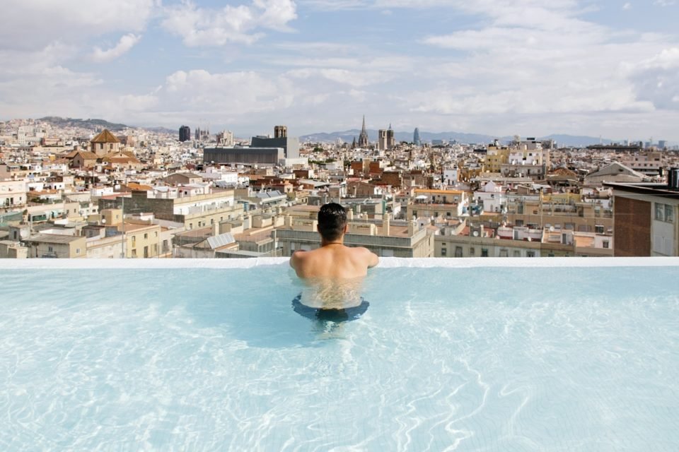 Um homem branco está de costas na beira da peiscina olhando para a cidade de Barcelona, na Espanha