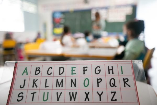 Foto colorida mostra uma sala de aula desfocada com uma placa de alfabeto em primeiro plano - Metrópoles