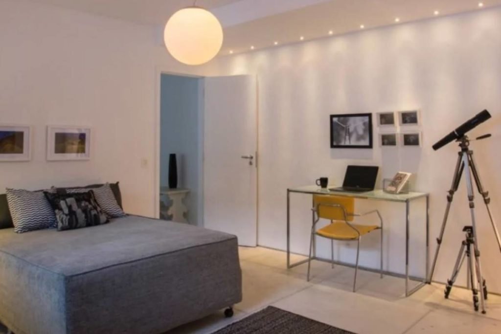 quarto com paredes brancas, cama com pano cinza e mesa com cadeira ao canto - metrópoles