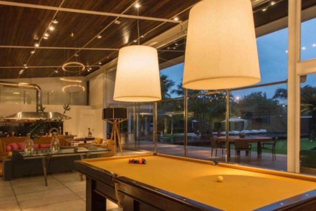 sala com mesa de sinuca, paredes de vidro e sofás ao fundo - metrópoles