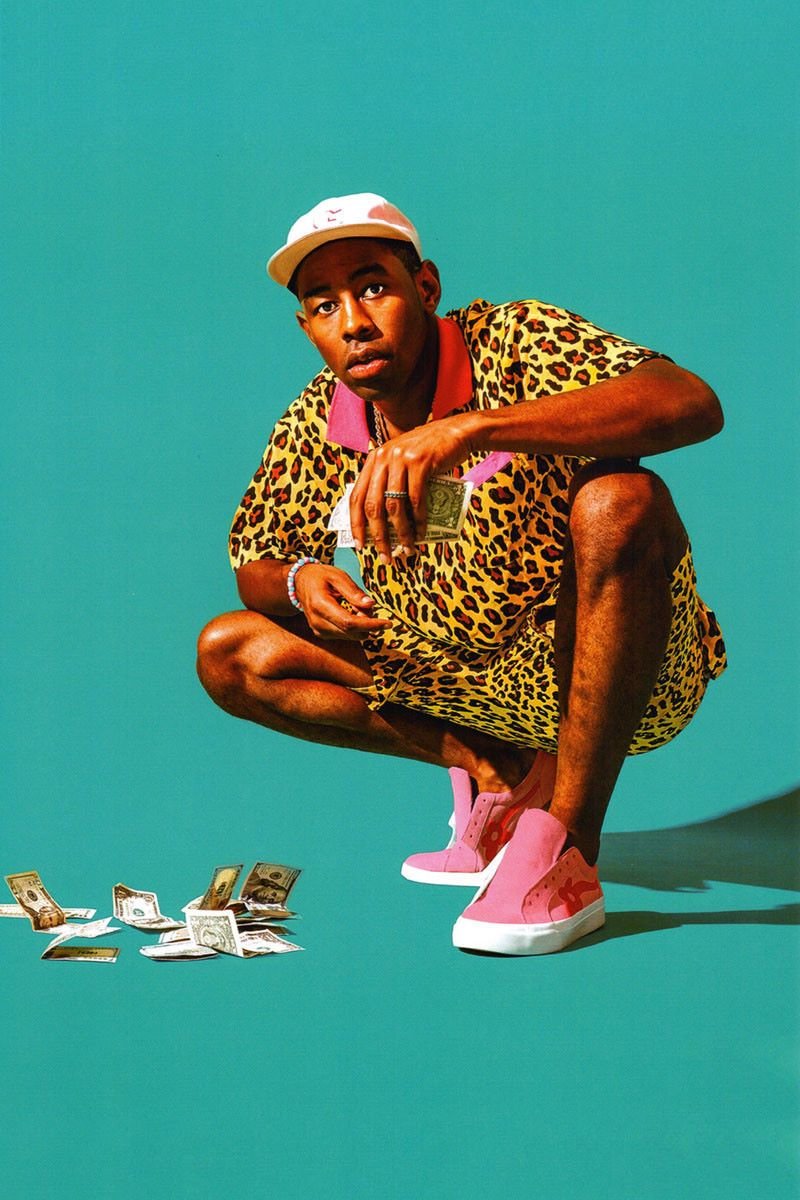 O rapper Tyler, The Creator, um homem jovem e negro, de cabelo raspado, posando para foto em um fundo azul. Ele usa um conjunto de camisa e bermuda, ambos com uma estampa de onça; um tênis rosa e um boné branco. As peças são da sua marca de moda, a Golf Wang. - Metrópoles