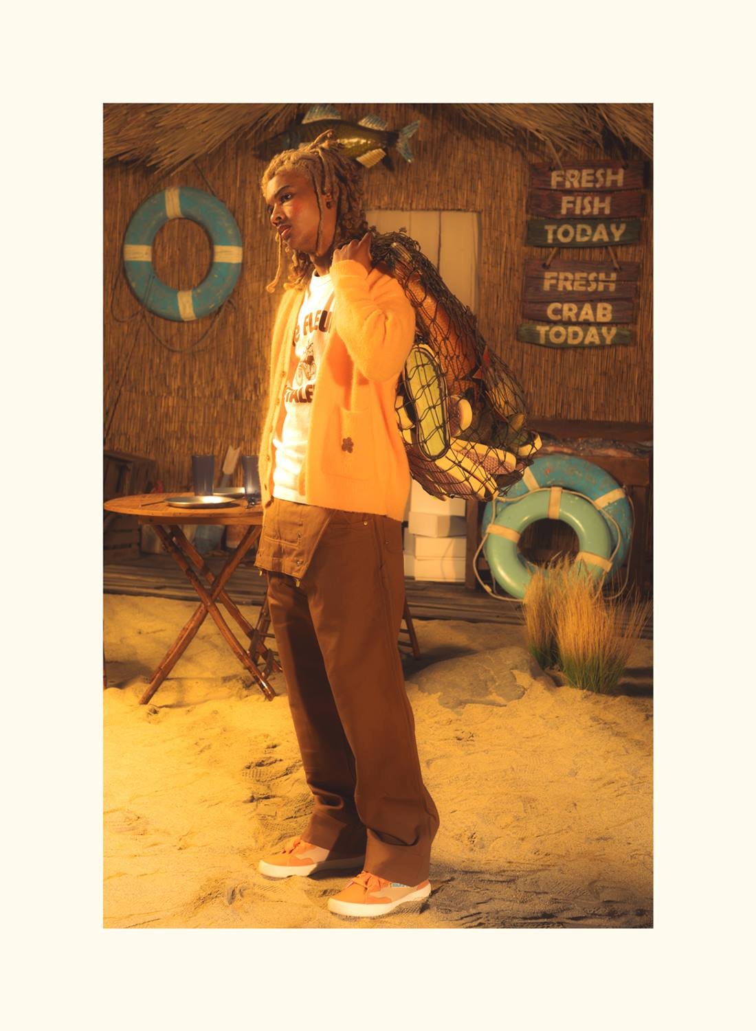 Modelo homem, jovem e negro, em campanha da marca Converse. Ele usa uma camiseta branca, uma calça marrom e um casaco laranja, além de um tênis colorido de camurça laranja. - Metrópoles