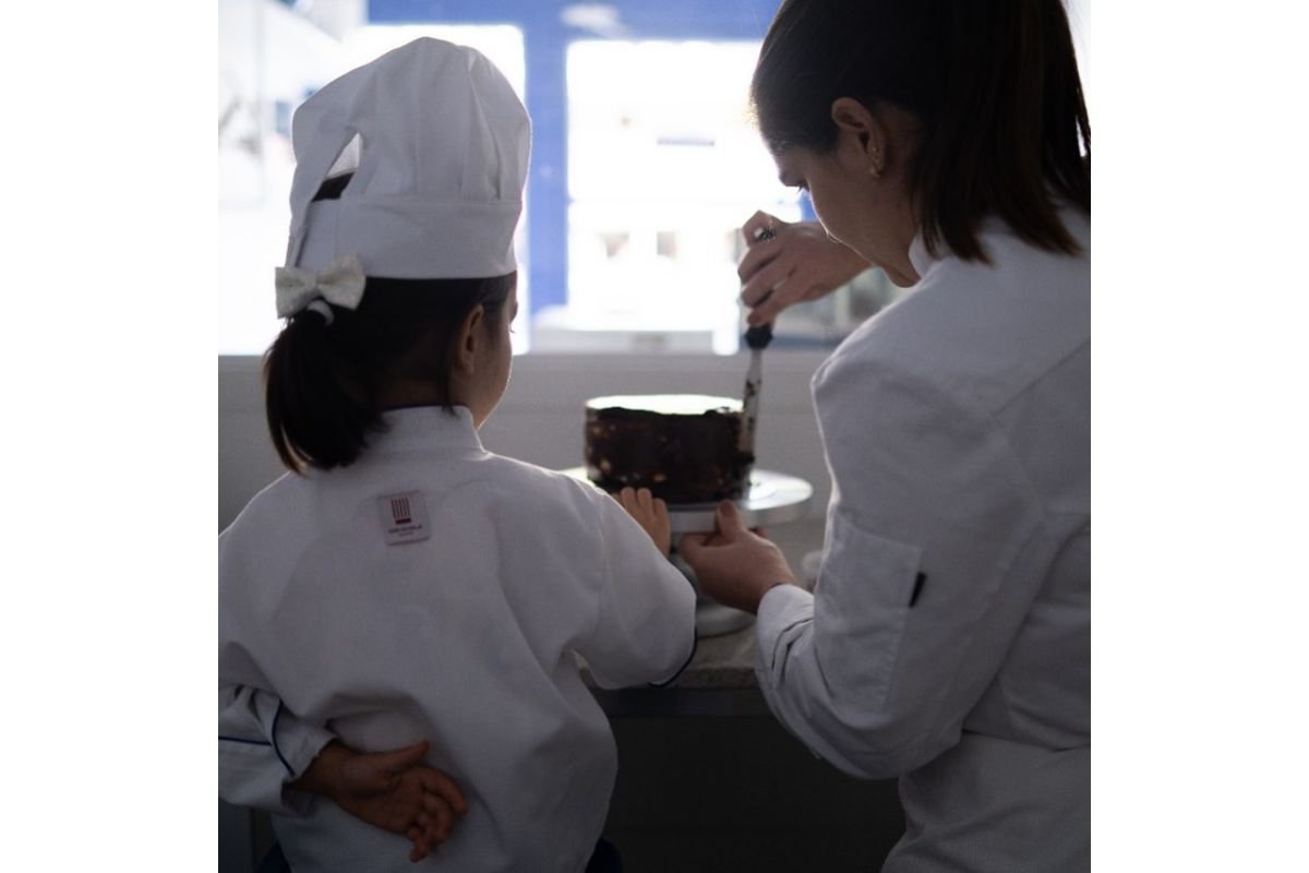 Foto colorida de menina e mulher de costas passando calda em um bolo de chocolate. Elas têm cabelos castanhos - Metrópoles