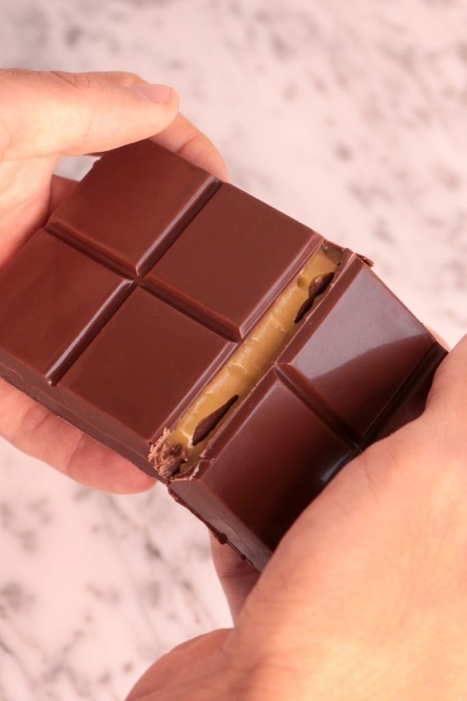 Foto colorida de mãos quebrando barra de chocolate recheada com doce de leite - Metrópoles