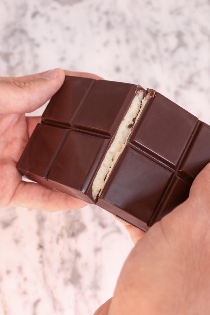 Foto colorida de mãos quebrando barra de chocolate com creme branco dentro - Metrópoles