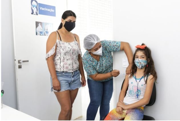 Imagem colorida de três pessoas: duas mulheres e uma criança. Uma das adultas está aplicando uma vacina no braço da criança