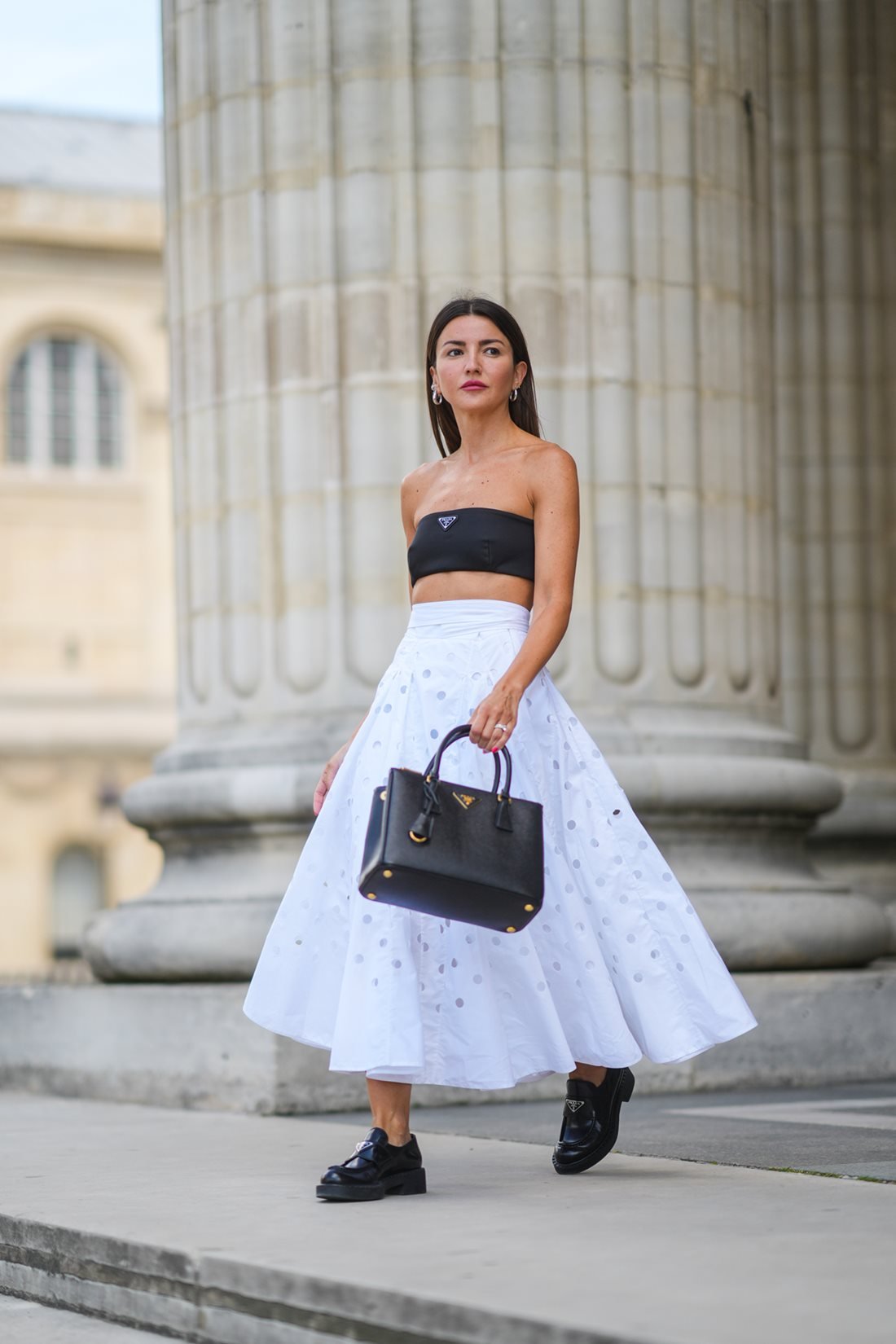 Young Caucasian woman with straight black hair walking down the street and posing for a photo.  She wears a black strapless top, a white skirt, black loafers and a leather bag called Galleria.  All pieces are by Prada.  - Metropolises