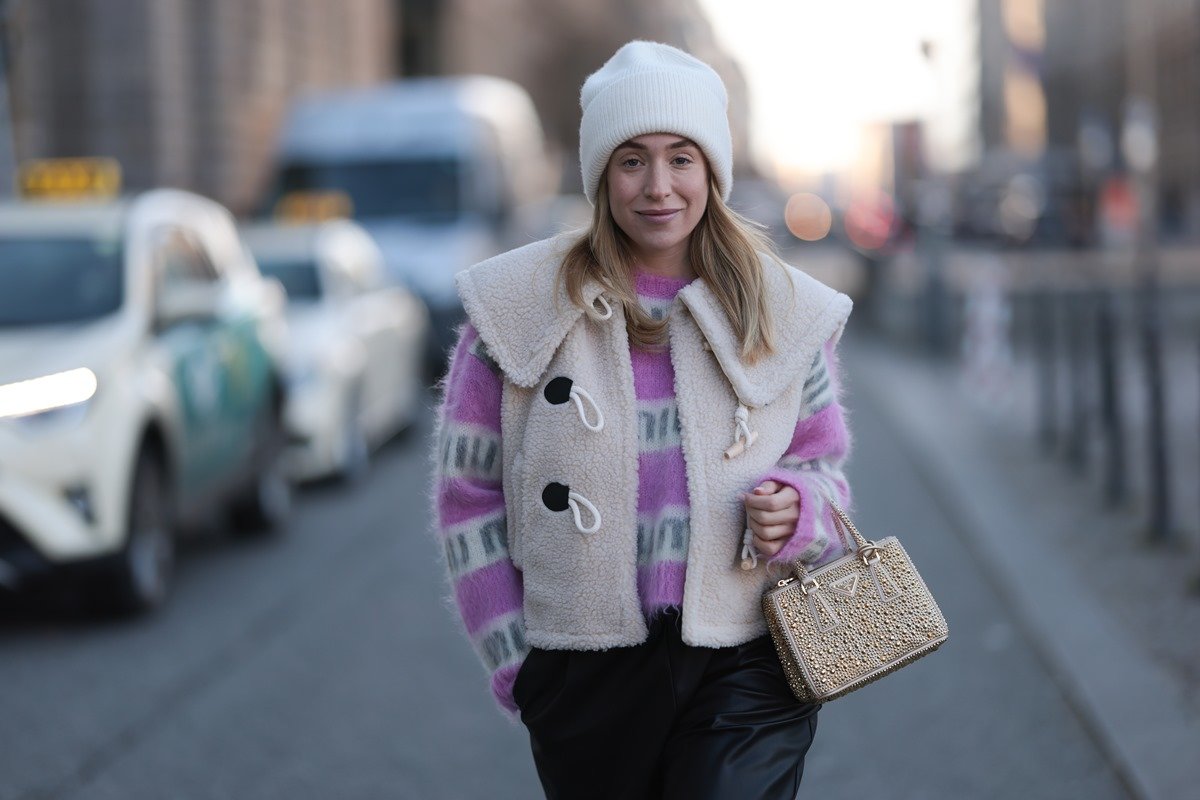 Young Caucasian woman with straight blonde hair walking on the street in Paris, France.  She wears a white and purple striped coat, a beige vest, a white cap, black leather pants and a beige bag, from the model Galleria, from the brand Prada.  - Metropolises