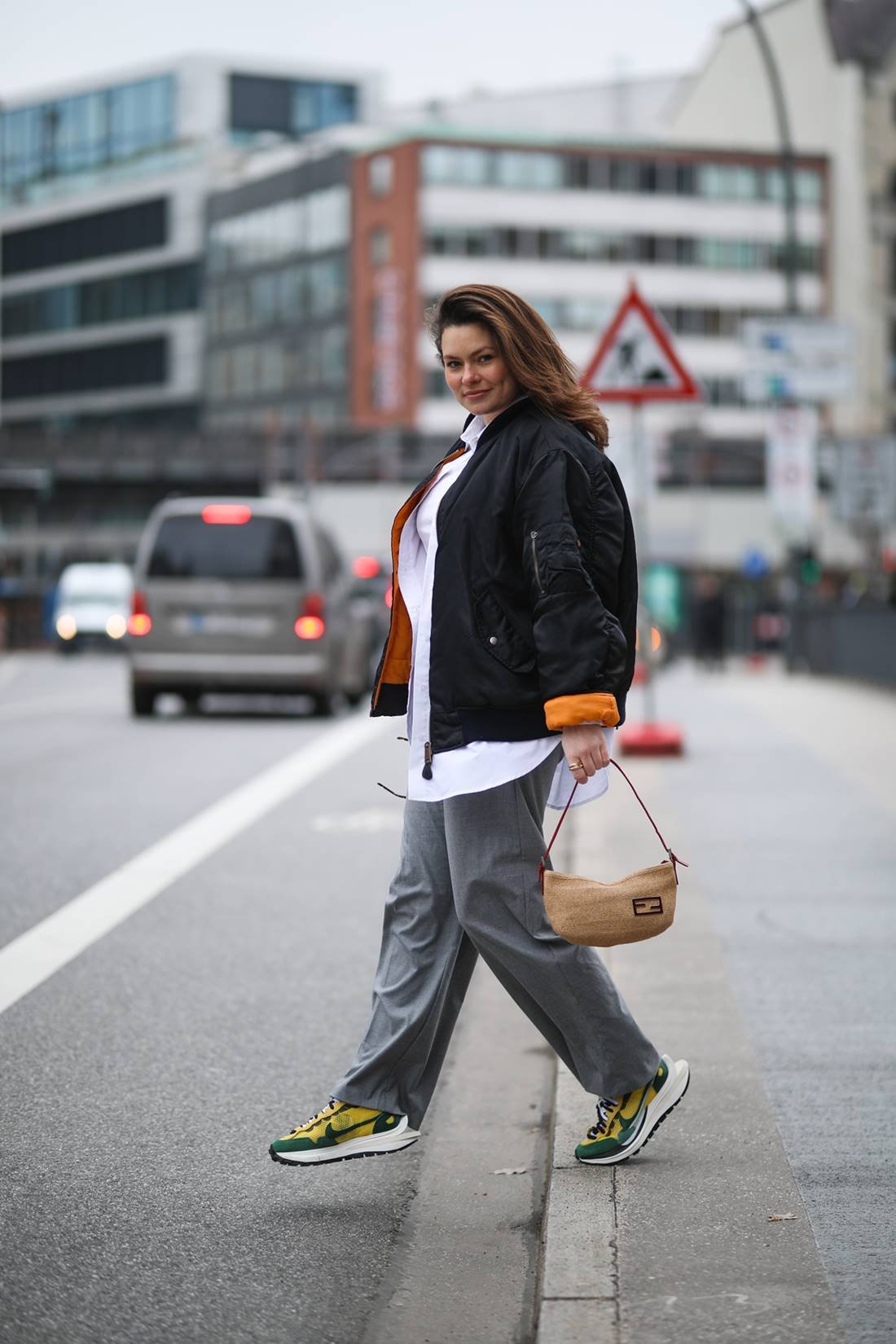 Young white woman with straight brown hair walking on the street.  He wears a white button-down shirt, baggy gray pants, Nike sneakers, and a black sports jacket.  She is holding a beige baguette bag by Fendi.  - Metropolises