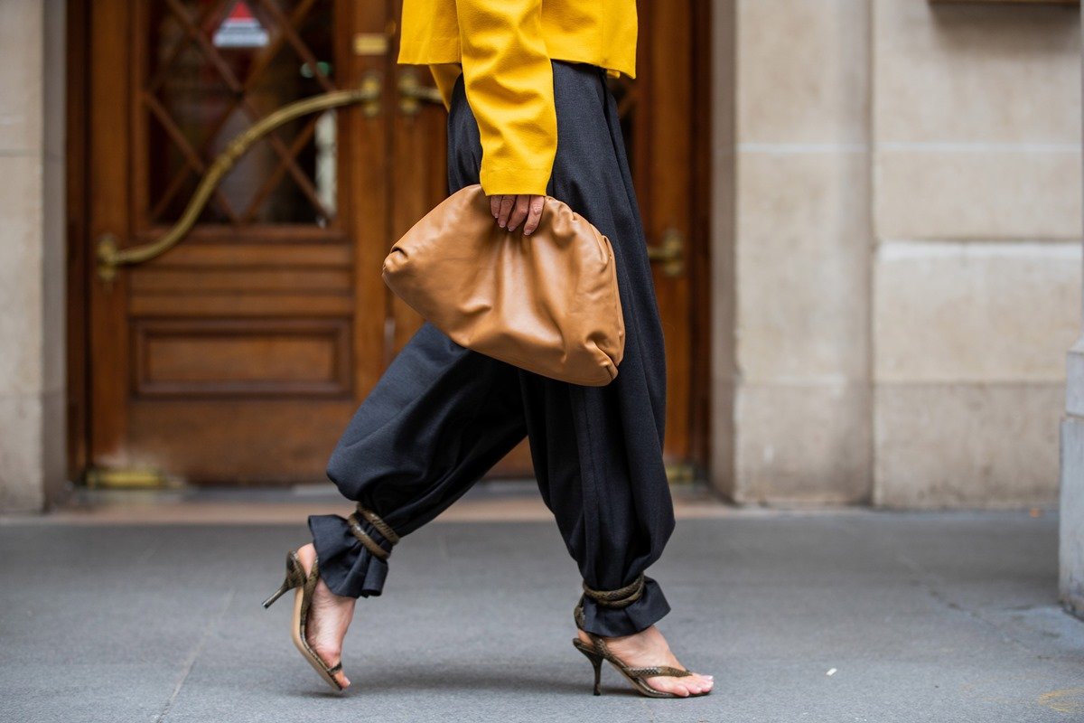 Young black woman walking on the street.  She wears baggy black trousers, a yellow coat, brown heeled sandals and a bag, model The Pouch, from the brand Bottega Veneta, in brown.  - Metropolises