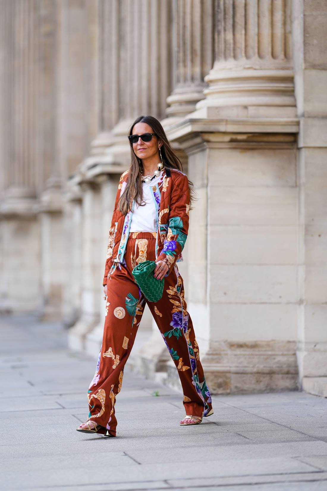 Middle aged Caucasian woman with long straight brown hair walking in the streets of Paris, France.  She wears a white t-shirt, a brown coat and pants set with colorful prints, a green leather bag, The Pouch model, from the Bottega Veneta brand, and Rayban sunglasses.  - Metropolises