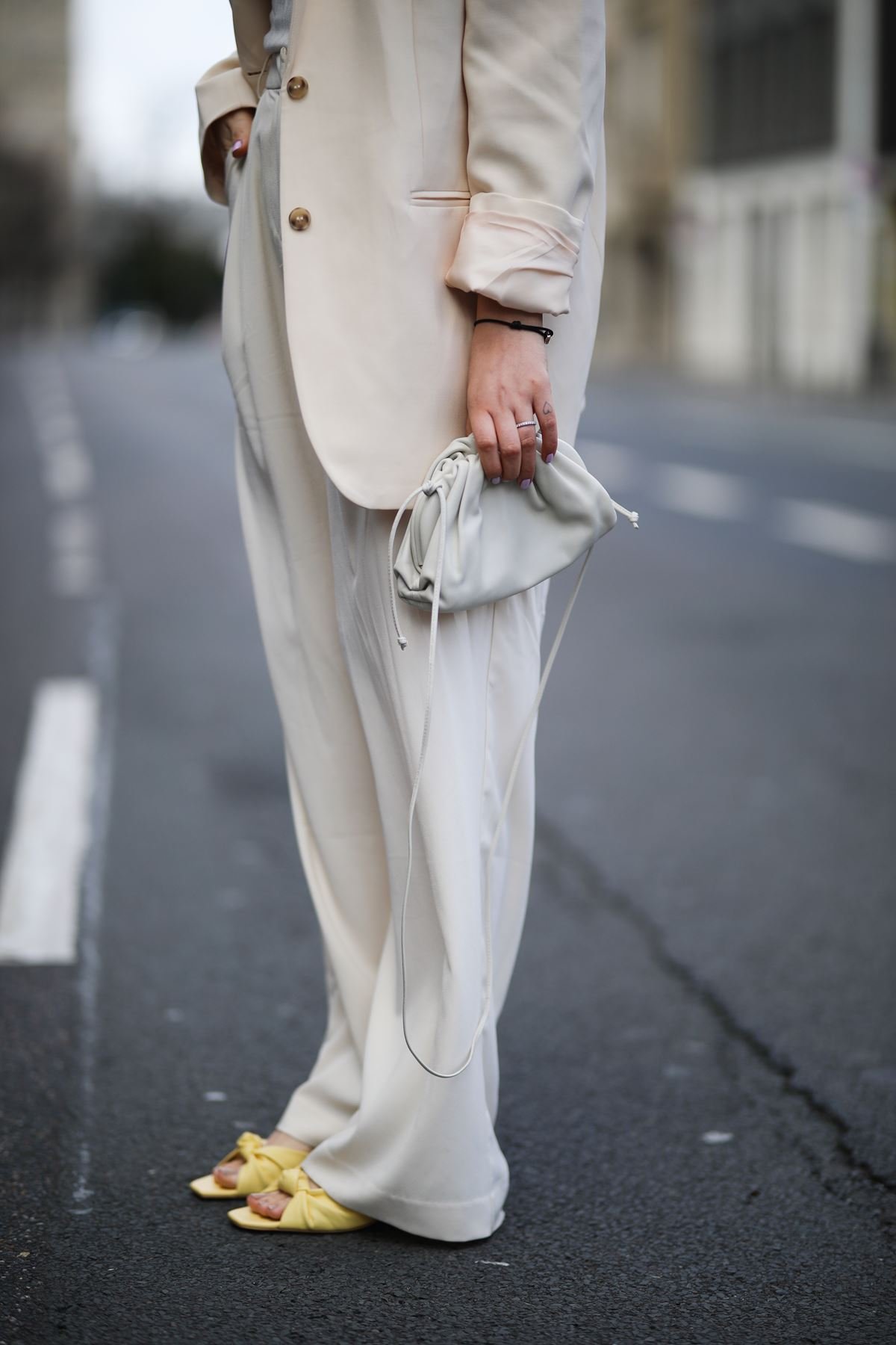 Photo of a person's legs during Paris Fashion Week.  The woman, who appears to be white and young, is wearing a cream turtleneck and pants set.  a yellow sandal with a heel;  and a bag called The Pouch by Bottega Veneta, also in cream.  - Metropolises