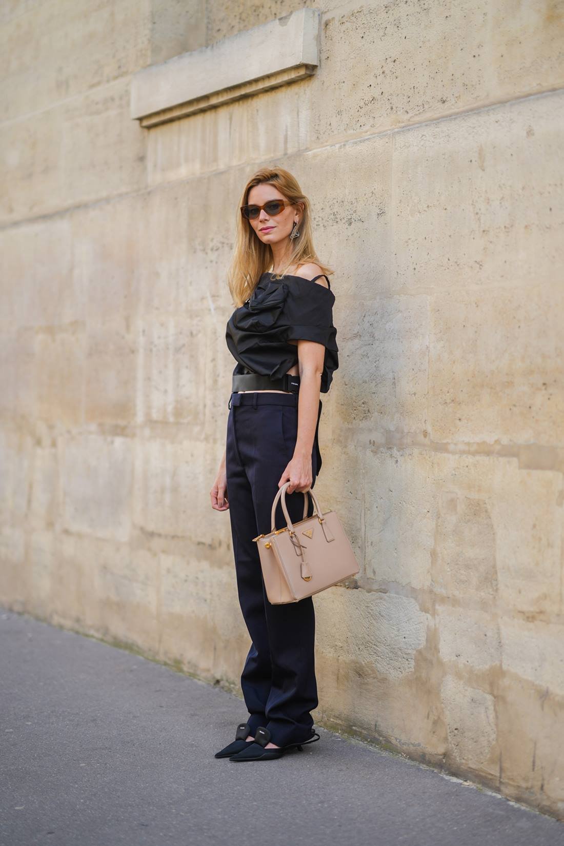 Young Caucasian woman with straight blond hair poses for a photo against a brown wall.  She wears a blouse and trousers, both in black, sunglasses and a beige leather Prada bag called Galleria.  - Metropolises