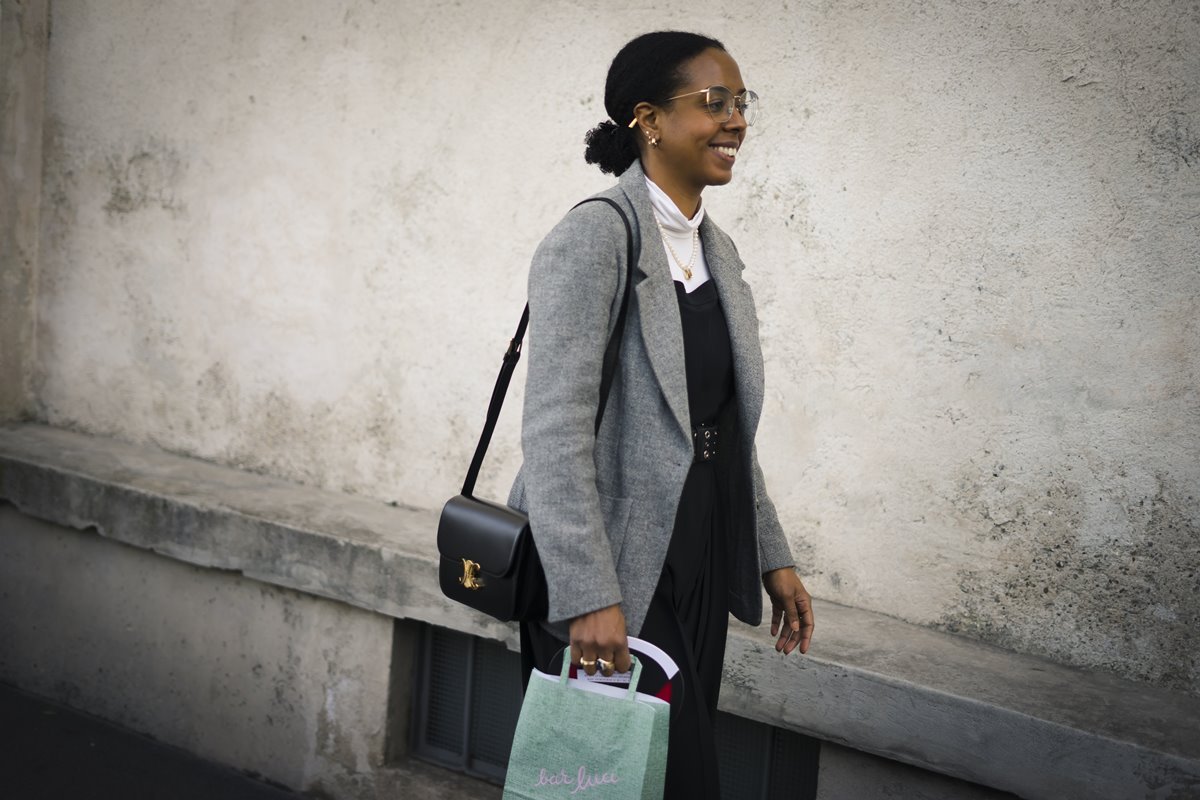 Mulher negra e jovem, de cabelo cacheado curto amarado em um coque, andando pela rua. Ela usa uma blusa de gola alta branca, um vestido preto por cima e um blazer cinza. - Metr&oacute;poles