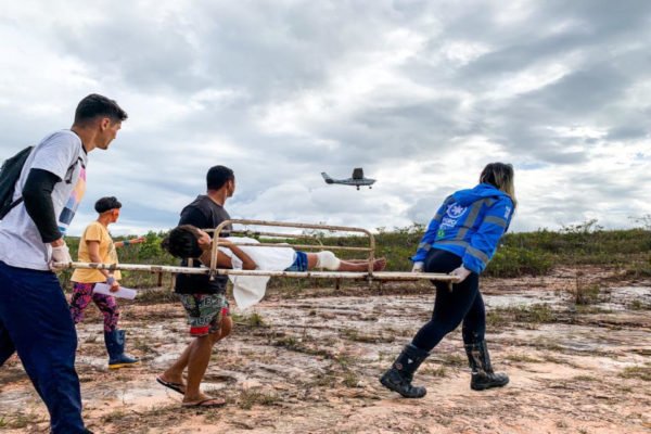 Voluntários da Associação Urihi Yanomami resgata criança indígena yanomani em maca vítima de desnetruição no norte do país. Ao fundo, é possível ver avião sobrevoando terreno. Crise dos Yanonamis - Metrópoles