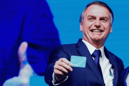 O ex-presidente Jair Bolsonaro mostra cartão à plateia em cerimônia do governo. Ele sorri, com telão atrás - Metrópoles