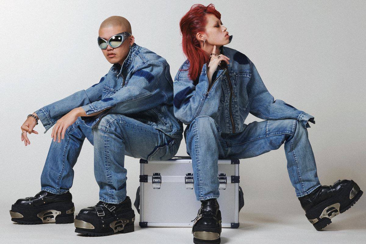 Dupla sendada em caixa prateada durante editorial de moda. Ambos estão usando peças jeans - Metrópoles
