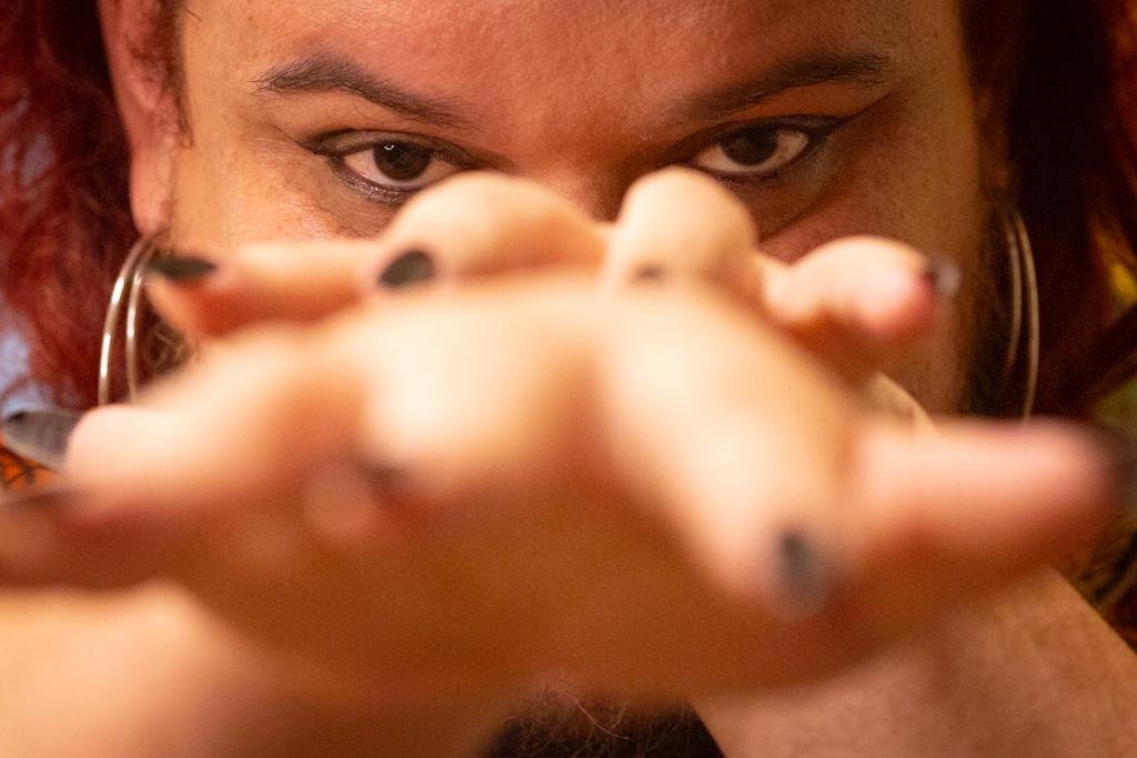 Mãos com unhas pretas encobrindo rosto de pessoa e mostrando apenas os olhos