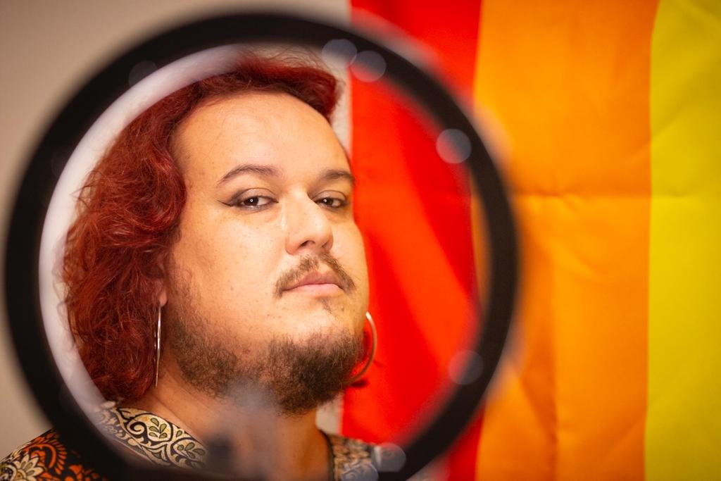 Pessoa com cabelo vermelho e barba olhando para foto. Ao fundo há uma bandeira vermelha e laranja