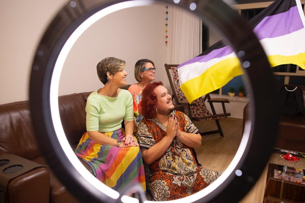 Pessoas sentadas em sala olhando para o lado. Há uma bandeira com as cores preta, roxa, branca e amarela ao lado