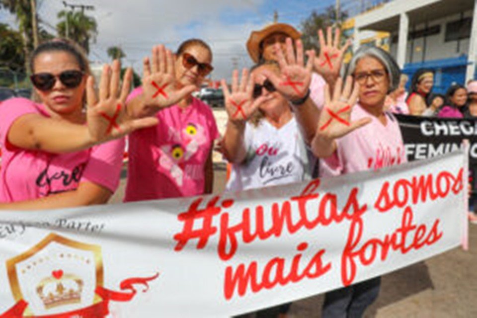 Mulheres de rosa mostrando a palma da mão com um x vermelho e segurando um cartaz
