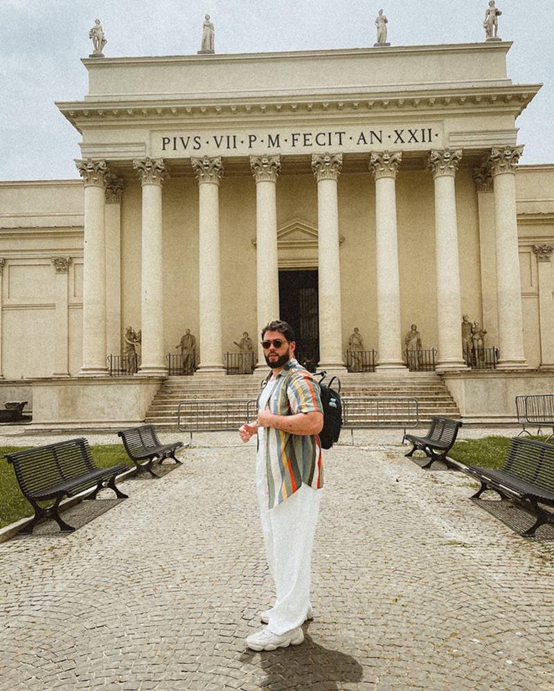 O influencer Vitor Arruda, um homem branco e jovem, de barba e cabelo preto, posa para foto em frente a um prédio do Vaticano, na Europa. Ele usa um camiseta branca; uma calça branca; uma camisa listrada colorida; uma mochila preta da marca Adidas. - Metrópoles