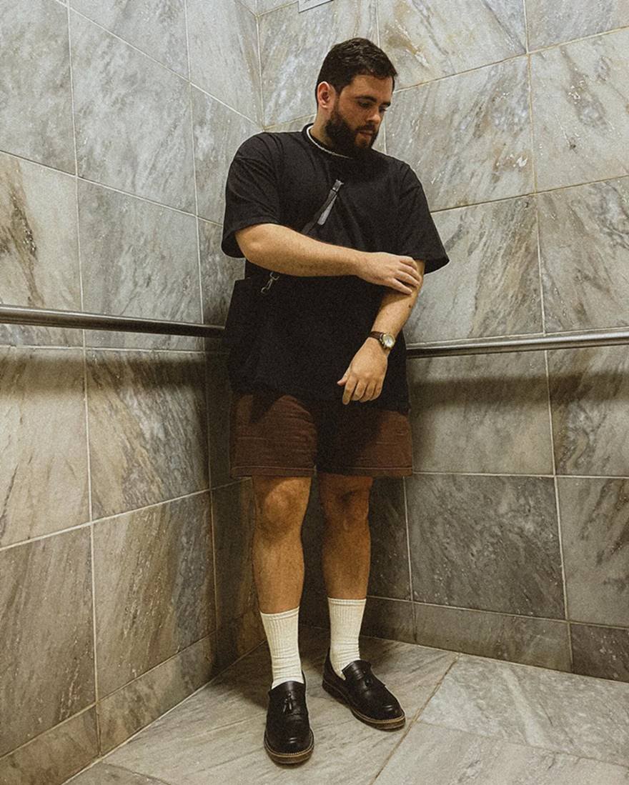 O influencer Vitor Arruda, um homem branco e jovem, de cabelo e barba preto, posa para foto em uma escadaria de marmoria. Usa uma camiseta preta, uma bermuda marrom, uma meia branca e um sapato mocassim de couro preto. - Metrópoles