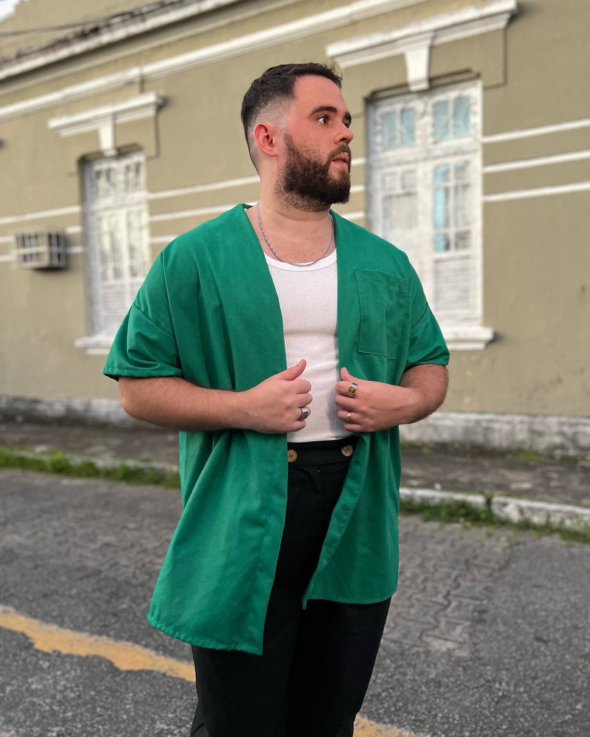 O influencer Vitor Arruda, um homem branco e jovem, de cabelo e barba preto, posa para foto na rua. Usa uma regata branca, uma camisa verde aberta por cima e uma calça preta. - Metrópoles