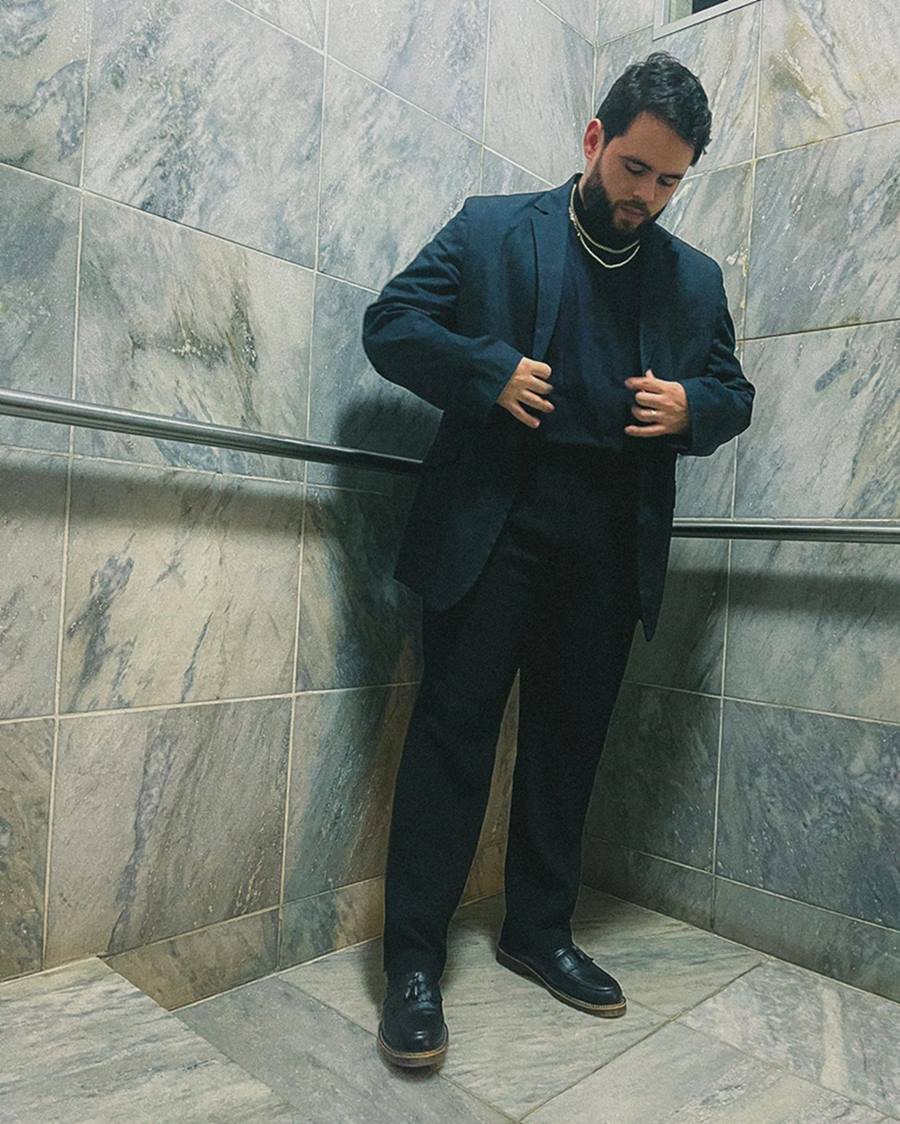 O influencer Vitor Arruda, um homem branco e jovem, de cabelo e barba preto, posa para foto em um corredor. Ele usa uma camiseta, um blazer e uma calça social, tudo na cor preta. - Metrópoles