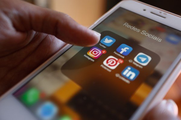 Fotografia colorida mostra pessoa apontando para pasta de aplicativos de redes sociais em celular