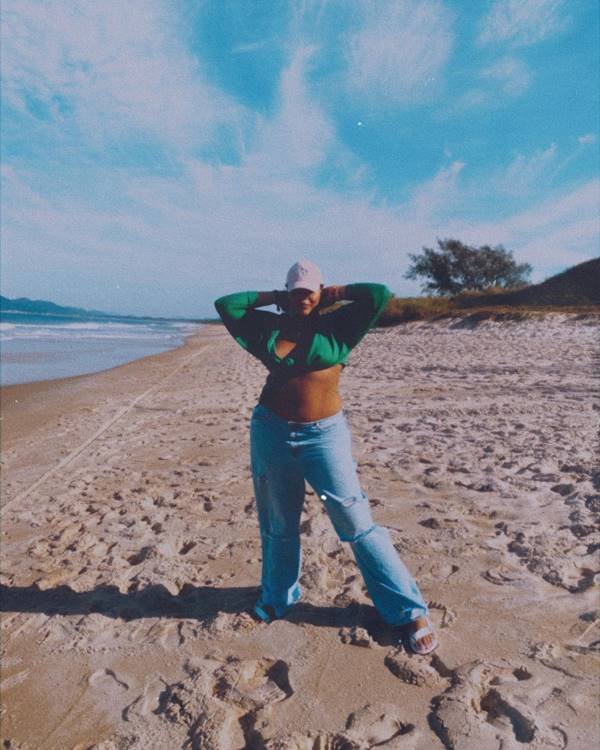 Modelo Rita Carreira na praia usa calça jeans de cintura baixa, top verde e boné - Metrópoles 