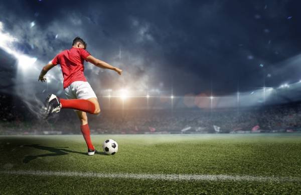 Imagem colorida mostrando jogador de futebol com camisa vermelha e bermuda branca se preparando para chutar uma bola em um estádio-Metrópoles