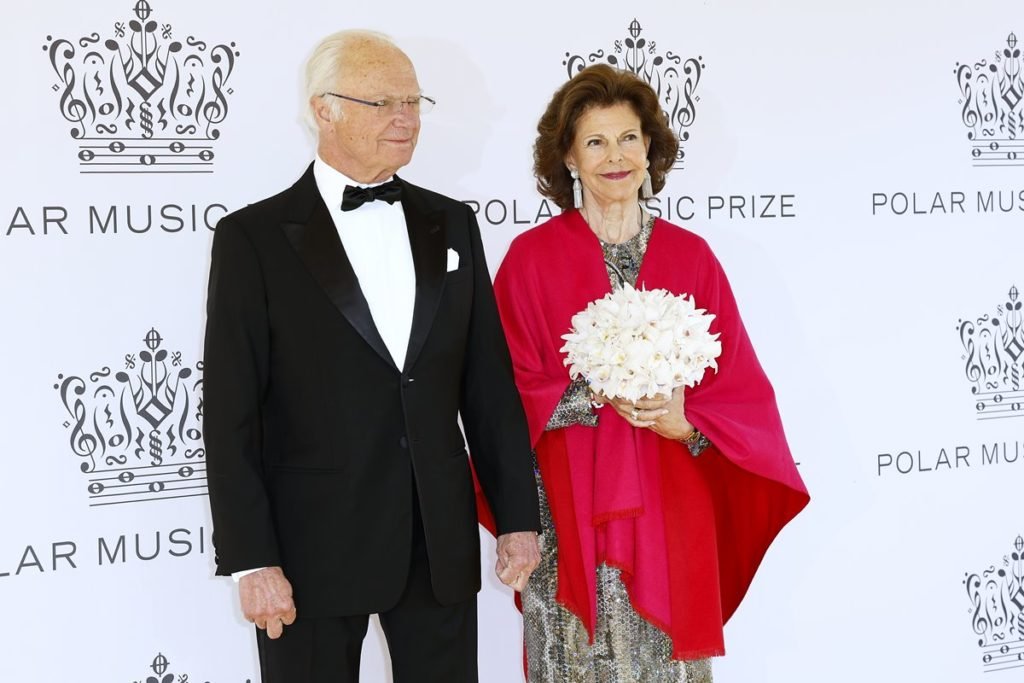 Foto colorida de homem branco, calvo, idoso e com fraque ao lado de mulher branca, idosa, com roupa vermelha e segurando buquê de flores - Metrópoles