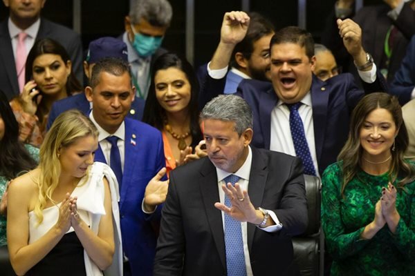 Imagem colorida de um homem de cabelos e barba grisalha ao lado de duas mulheres e ao fundo alguns homens usando terno e gravata