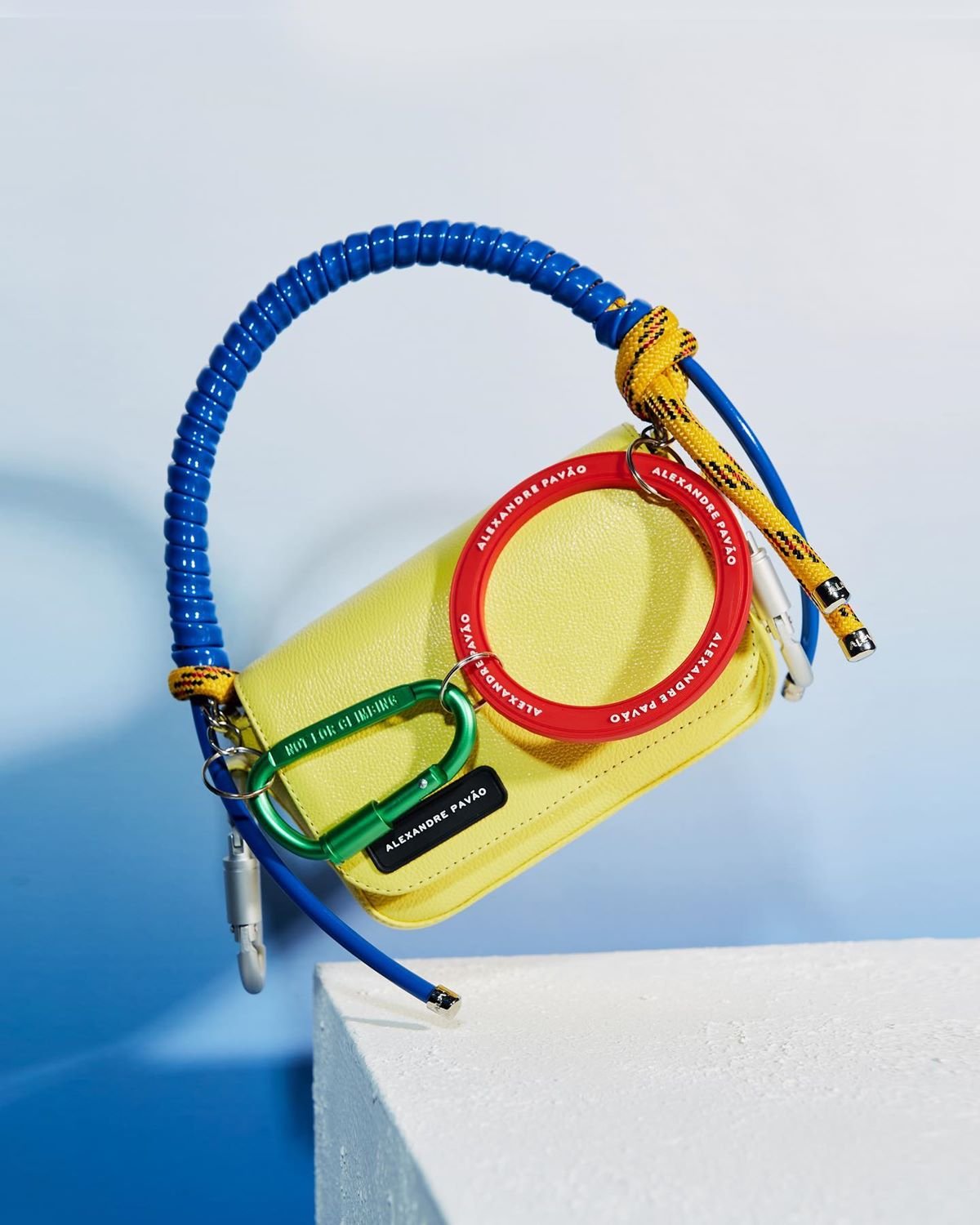 Bolsa retangular e de couro amarelo, com alça transpassada azul e dois chaveios de ferro nas cores vermelha e verde. A peça é da marca Alexandre Pavão. - Metrópoles