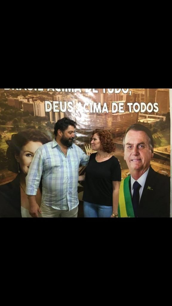 João Baldan, preso na Operação Lesa Pátria por participar de atos antidemocráticos, e a deputada federal Carla Zambelli (PL-SP). Eles se abraçam, diante de um poster com fotos de campanha de Zambelli e Bolsonaro - Metrópoles