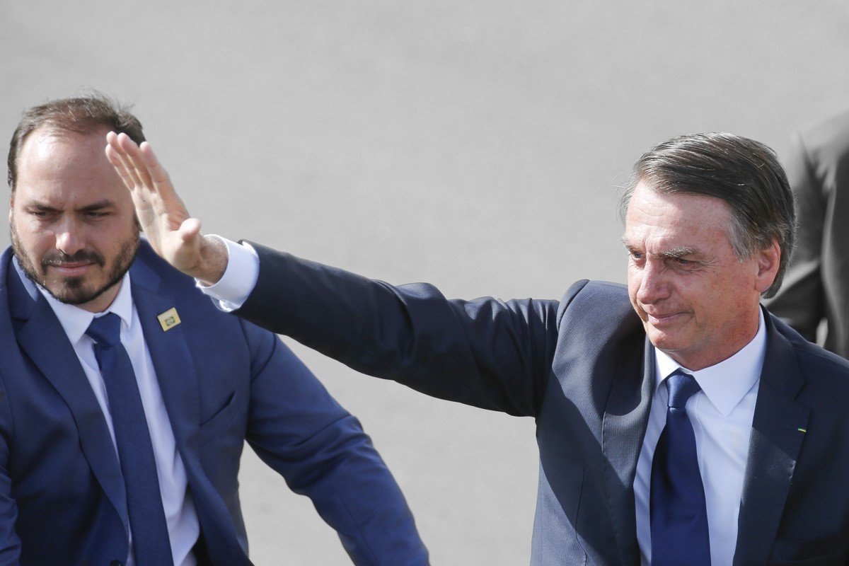 O ex-presidente Bolsonaro acena em evento ao lado de seu filho, Carlos Bolsonaro. Ambos sorriem e usam terno - Metrópoles
