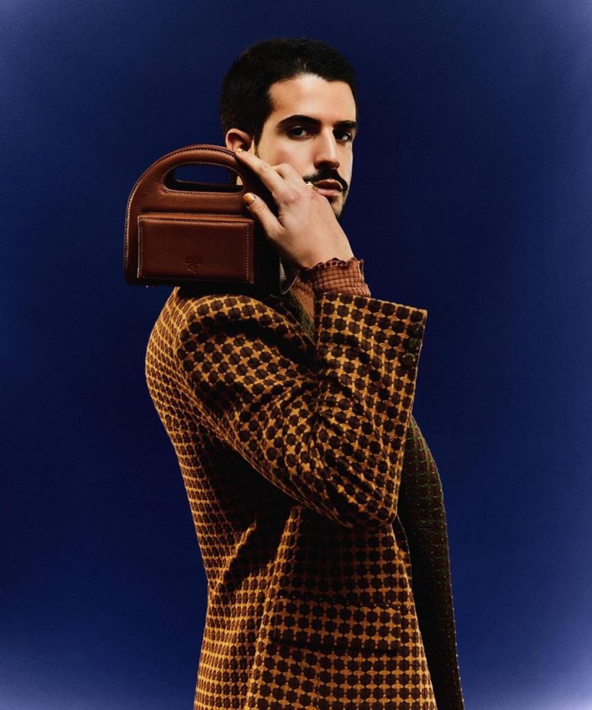 Enzo Cellolari poses for a photo against a simple blue background.  He is a white young man with black hair and a mustache.  He wears a brown checked jacket and a brown leather bag.  All fashion items are from Misci brand.  - capital Cities