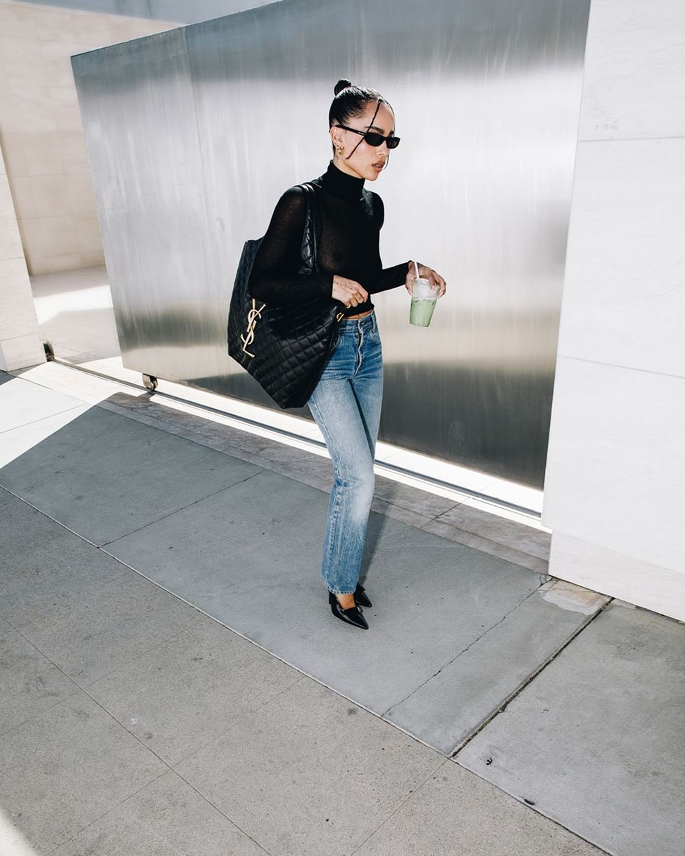 A atriz Zoë Kravitz, uma mulher negra e jovem, de cabelo preto liso, posa para campanha da marca Saint Laurent enquanto anda na rua. Ela usa uma blusa preta, de manga longa, uma calça jeans, óculos escuros e uma bolsa preta, grande, modelo Icare. - Metrópoles