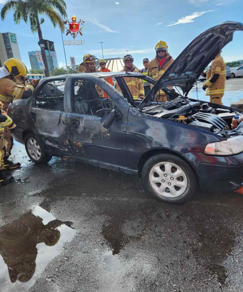 Lateral de Fiat Siena azul com o capô aberto. Carro foi consumido pelas chamas. Foto colorida. Vista diagonal dianteira do veículo