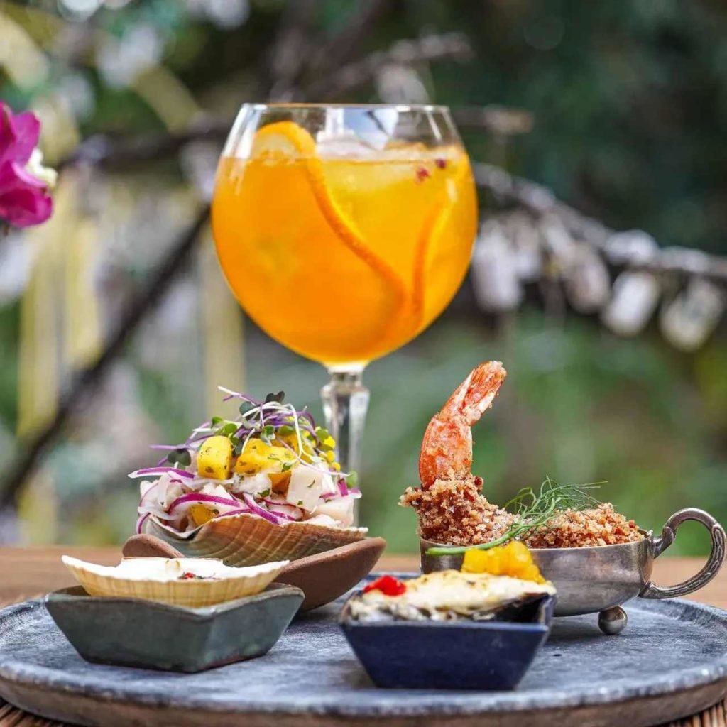 Na foto, vários pratos com comidas e uma taça com drinque laranja - Metrópoles